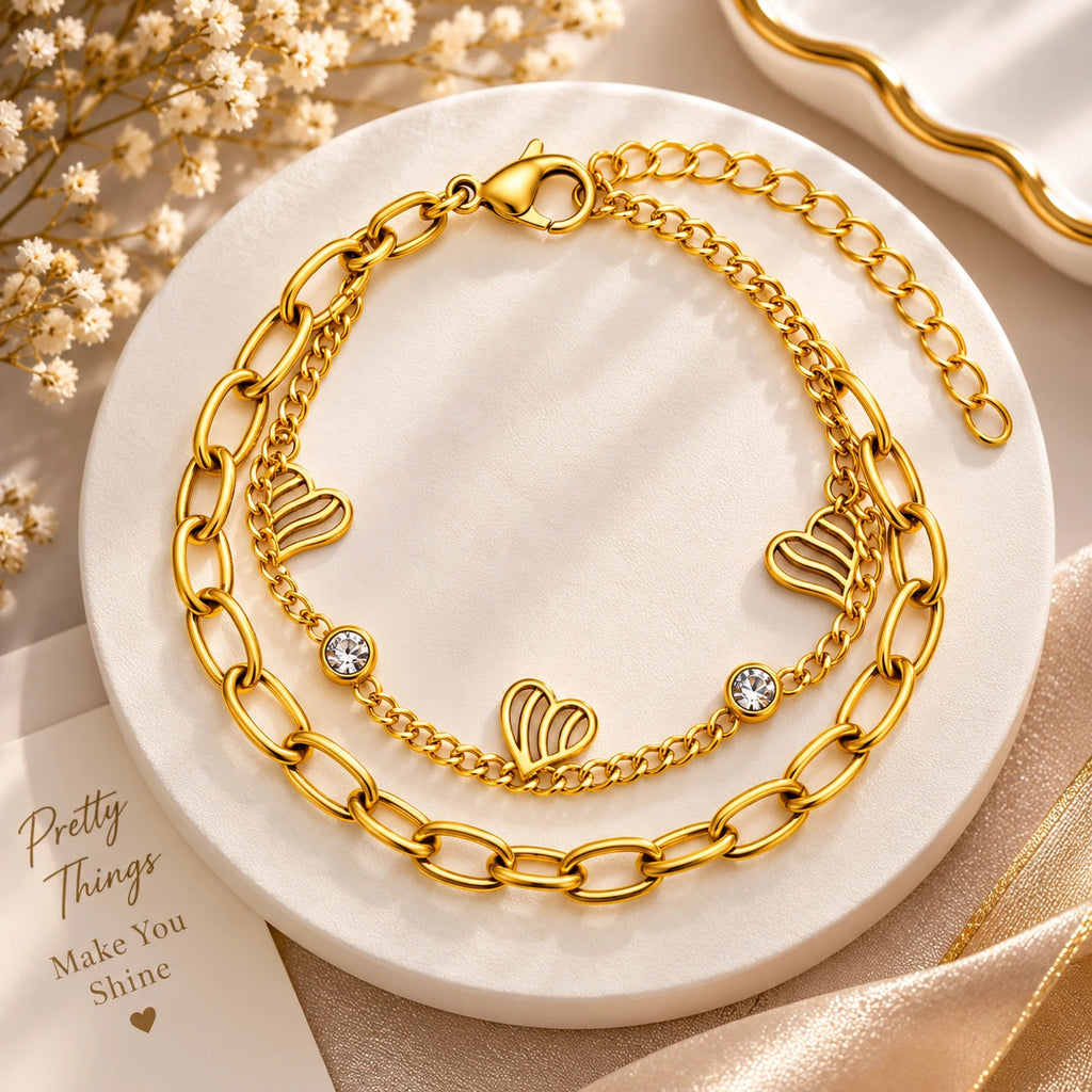 A gold-toned double-layer bracelet displayed on a round white marble tray. The top layer features a chunky paperclip chain, and the bottom layer is a delicate chain with three open heart charms and two small clear crystal accents.
