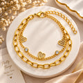 A gold-toned double-layer bracelet displayed on a round white marble tray. The top layer features a chunky paperclip chain, and the bottom layer is a delicate chain with three open heart charms and two small clear crystal accents.