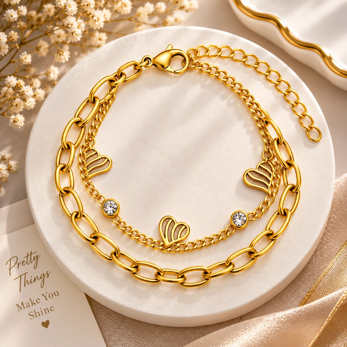 A gold-toned double-layer bracelet displayed on a round white marble tray. The top layer features a chunky paperclip chain, and the bottom layer is a delicate chain with three open heart charms and two small clear crystal accents.