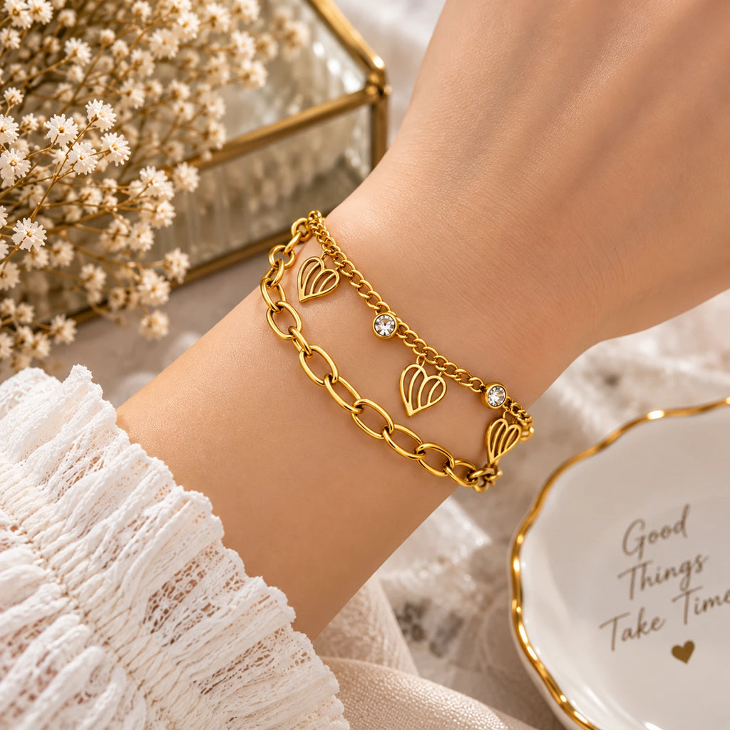 A gold-toned double-strand bracelet with heart-shaped links and small crystal accents, worn on a person's wrist. The scene includes a white lace sleeve, dried flowers, a gold-framed mirror, and a white dish with gold lettering.