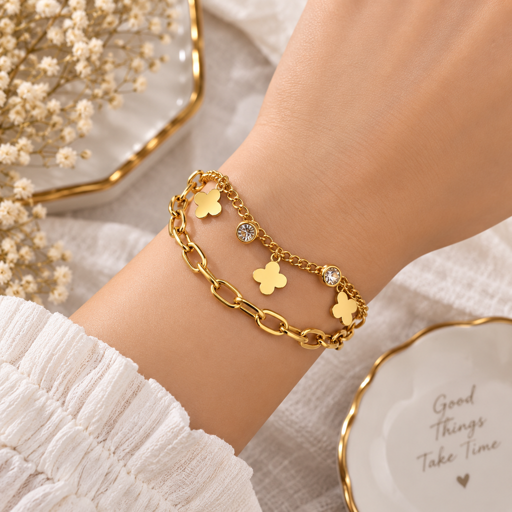 A close-up of a person's wrist wearing a gold chain bracelet with four clover-shaped charms, two of which are adorned with clear rhinestones. The background features a white textured fabric, dried baby's breath in a gold dish, and a white plate with gold trim.