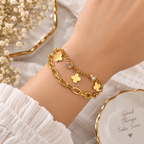 A close-up of a person's wrist wearing a gold chain bracelet with four clover-shaped charms, two of which are adorned with clear rhinestones. The background features a white textured fabric, dried baby's breath in a gold dish, and a white plate with gold trim.