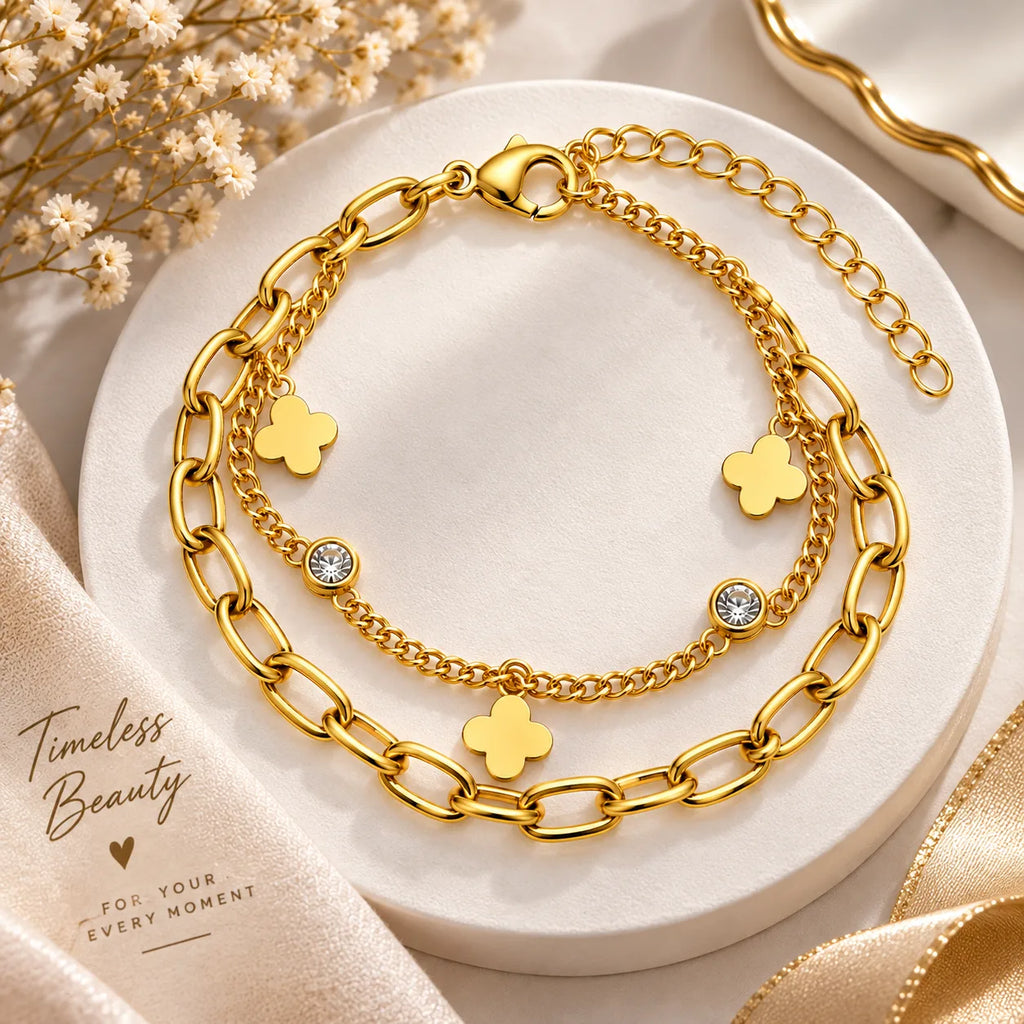A layered gold-tone chain bracelet set displayed on a round white pedestal. The set includes a thick paperclip chain, a delicate chain, and three small gold clover charms, with two charms featuring clear rhinestones.