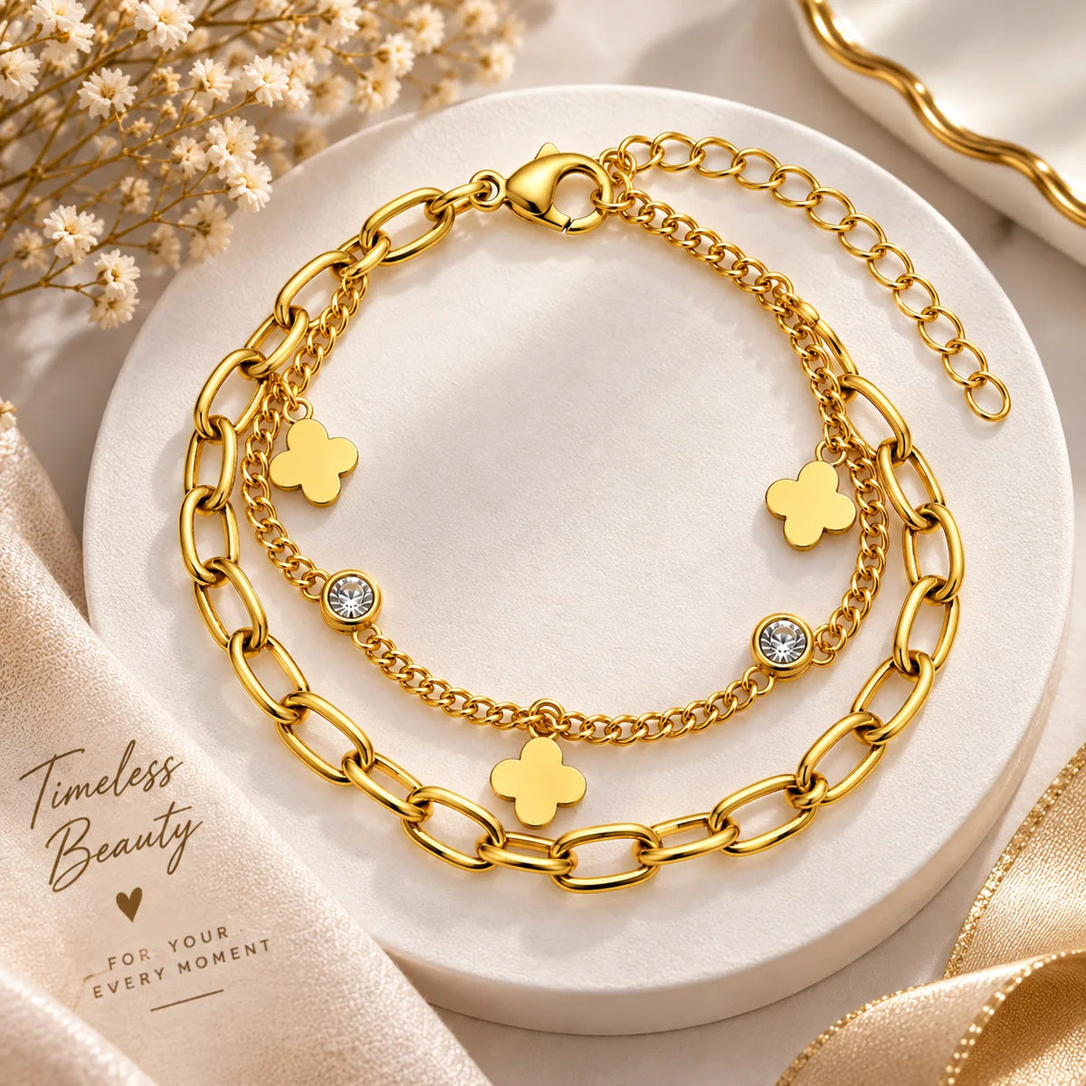 A layered gold-tone chain bracelet set displayed on a round white pedestal. The set includes a thick paperclip chain, a delicate chain, and three small gold clover charms, with two charms featuring clear rhinestones.