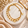 A gold-toned layered chain bracelet with three dragonfly charms, two small crystal-set round charms, and an extender chain, displayed on a round white pedestal with decorative dried flowers and satin fabric.