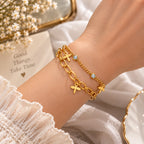 A close-up of a person's wrist wearing two delicate gold bracelets, one with an elongated oval link design and one with a twisted chain and small dragonfly charms, set against a soft white fabric background with baby's breath flowers.