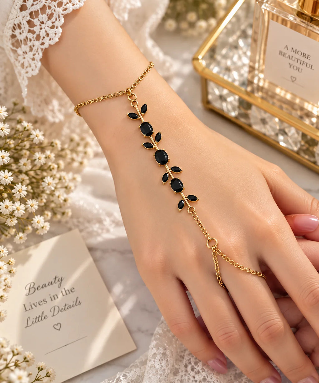 A gold leaf vine hand chain bracelet with black oval stones, worn on a woman's wrist. The image is styled with a lace sleeve, white baby's breath flowers, and a gold-framed perfume bottle in the background.