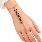 A gold chain hand bracelet worn on a woman's wrist and fingers, featuring a leafy vine design with black oval and round stones, shown against a white background.