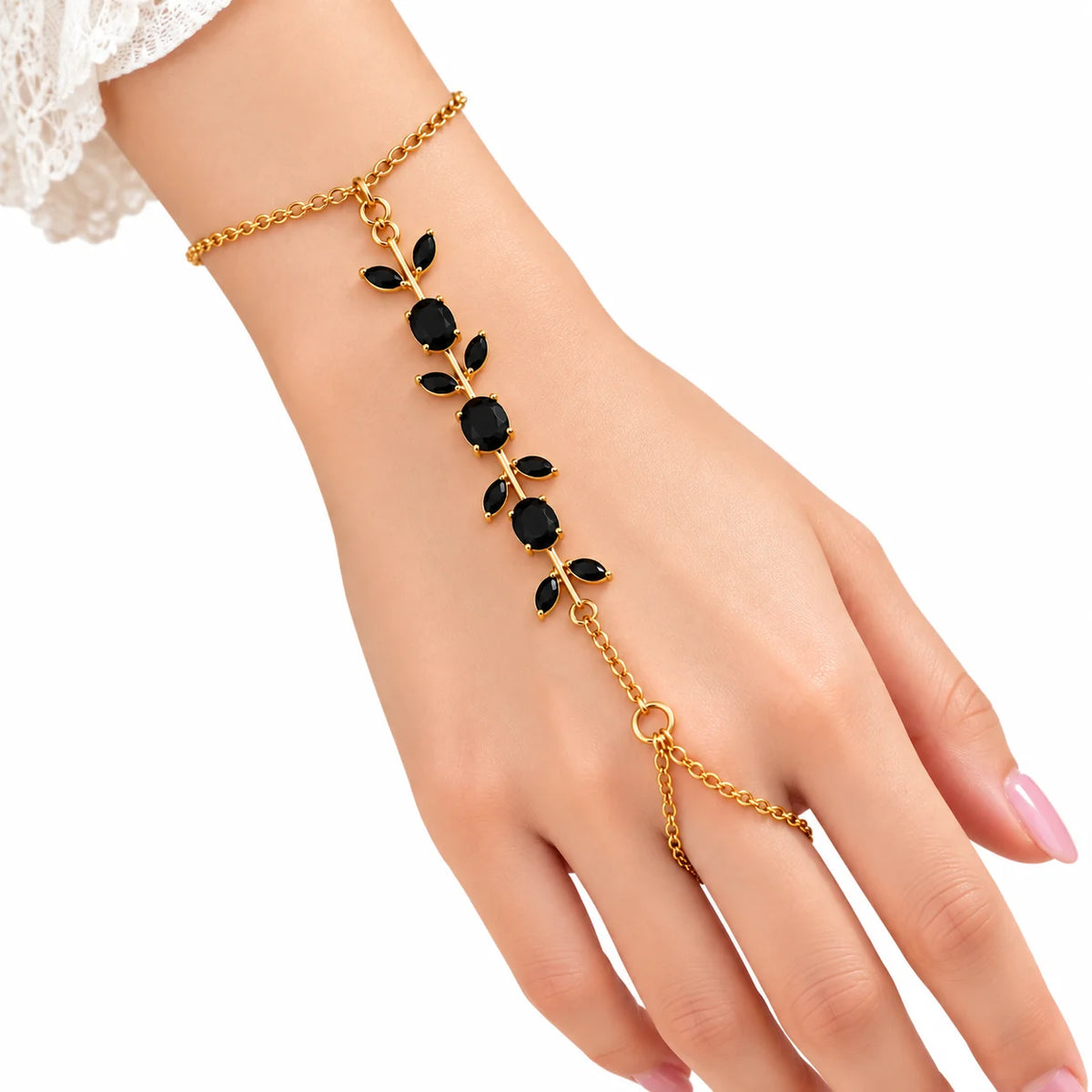 A gold chain hand bracelet worn on a woman's wrist and fingers, featuring a leafy vine design with black oval and round stones, shown against a white background.