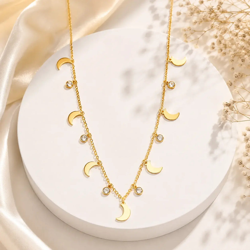 A gold-toned double-strand necklace with moon phase charms and small clear crystal accents, displayed on a white round pedestal with soft satin and dried flowers in the background.