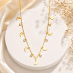A gold-toned double-strand necklace with moon phase charms and small clear crystal accents, displayed on a white round pedestal with soft satin and dried flowers in the background.