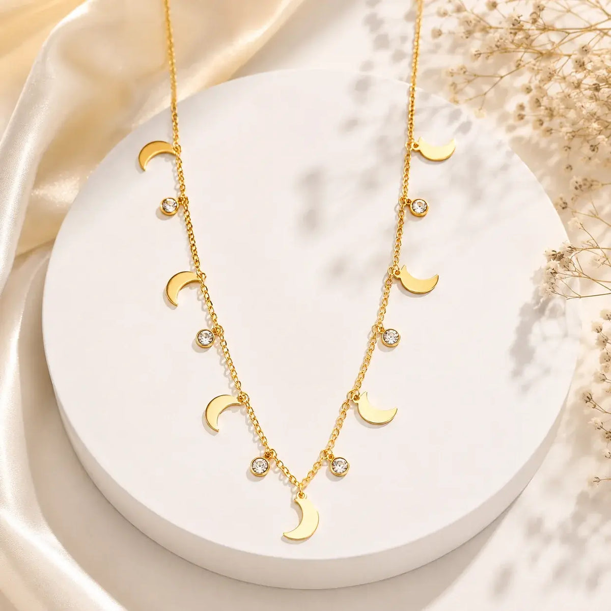 A gold-toned double-strand necklace with moon phase charms and small clear crystal accents, displayed on a white round pedestal with soft satin and dried flowers in the background.