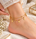 A close-up of a person's ankle and foot wearing a delicate gold double-layer anklet with star charms, styled with lace-trimmed socks and small white flowers.