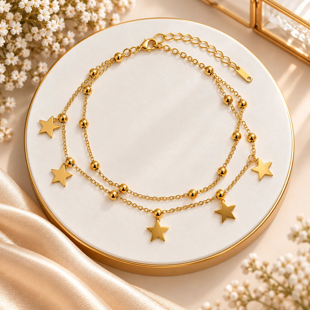 A gold-toned double-strand chain necklace with five star charms and small bead accents, displayed on a round white and gold tray with satin fabric and baby's breath flowers.