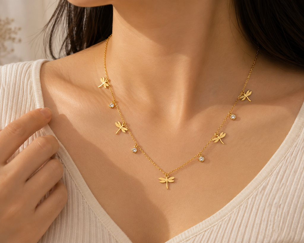 A close-up of a woman wearing a delicate gold necklace with five dragonfly charms and small clear stone accents, styled with a cream ribbed top.