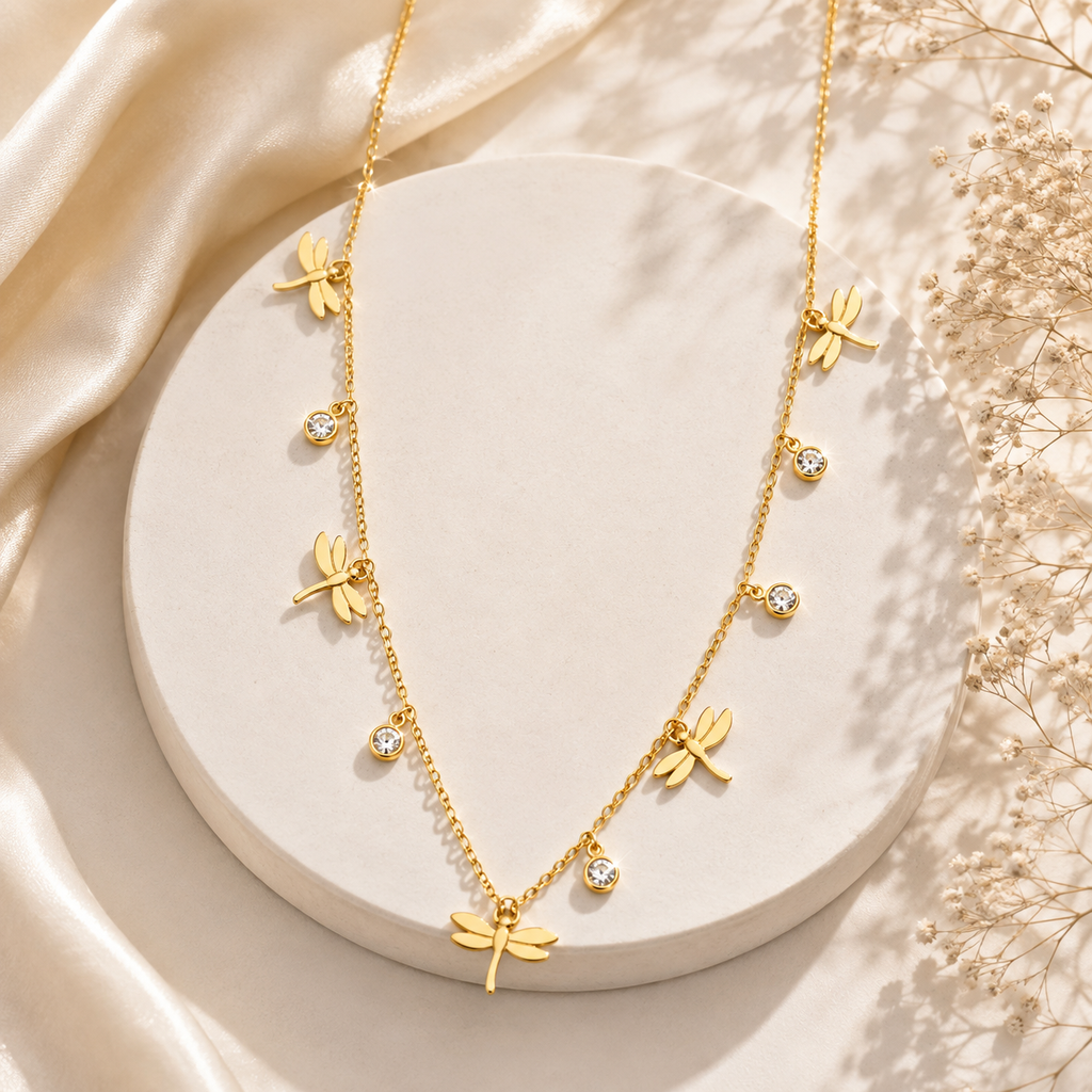 A gold necklace with dragonfly charms and small clear crystal accents, displayed on a round cream pedestal with draped satin and dried flowers in the background.