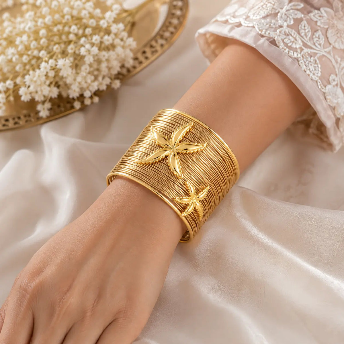 A wide gold cuff bracelet with two raised starfish motifs, worn on a person's wrist over white lace fabric, with a decorative tray of white flowers in the background.