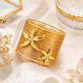A wide gold cuff bracelet with two raised starfish motifs, styled on a soft cream satin surface with dried flowers and decorative props.