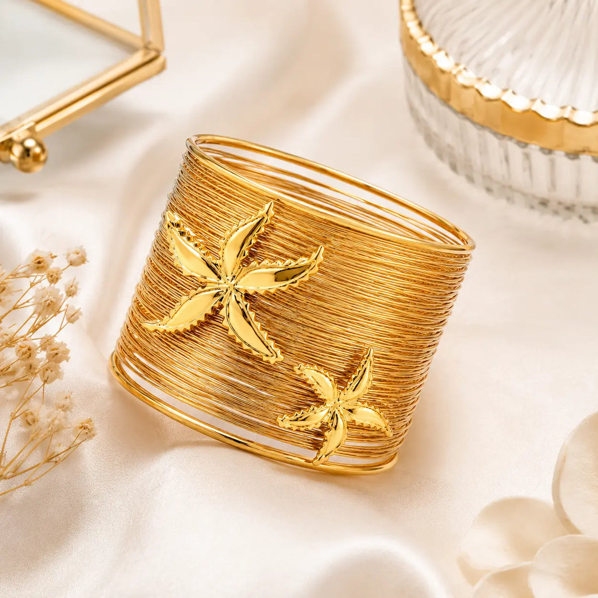 A wide gold cuff bracelet with two raised starfish motifs, styled on a soft cream satin surface with dried flowers and decorative props.