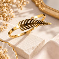 A polished gold bangle bracelet shaped like a fern frond, resting on a textured cream stone block with dried flowers and soft neutral fabric in the background.