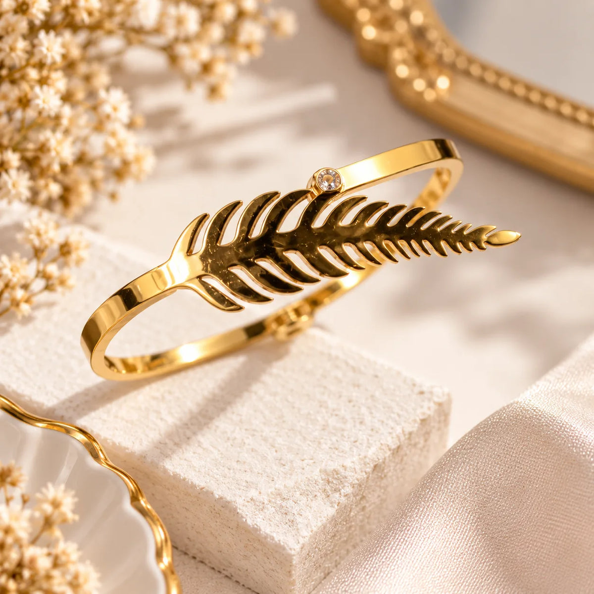 A polished gold bangle bracelet shaped like a fern frond, resting on a textured cream stone block with dried flowers and soft neutral fabric in the background.