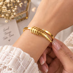 A gold leaf-shaped cuff bracelet with a small clear crystal accent, shown worn on a person's wrist with manicured nails visible, set against a soft white background with decorative props.