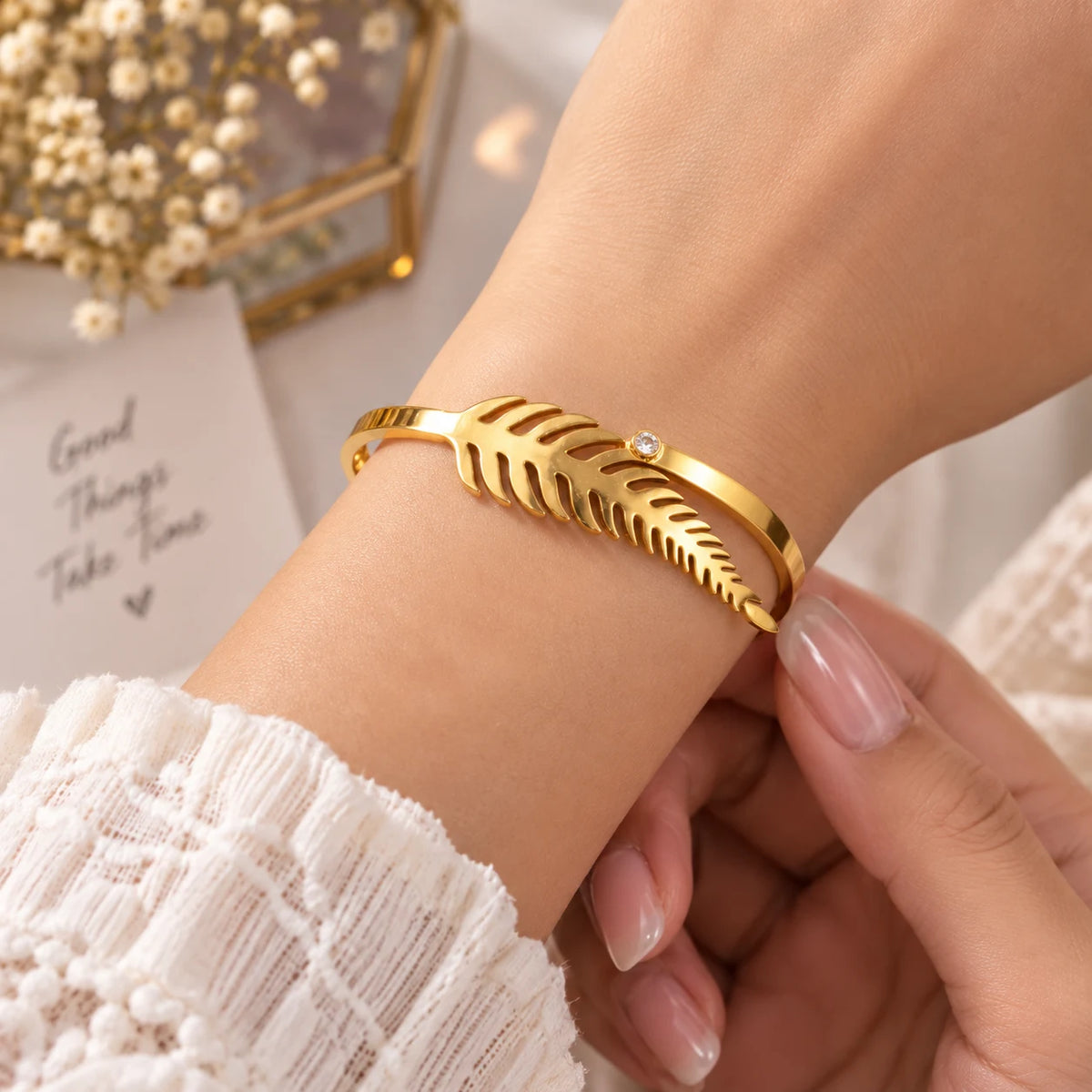 A gold leaf-shaped cuff bracelet with a small clear crystal accent, shown worn on a person's wrist with manicured nails visible, set against a soft white background with decorative props.