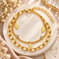 A gold-toned double-layer bracelet with chunky oval links, delicate chain links, leaf charms, and crystal-studded accents, displayed on a round white marble tray with dried flowers and soft beige fabric.