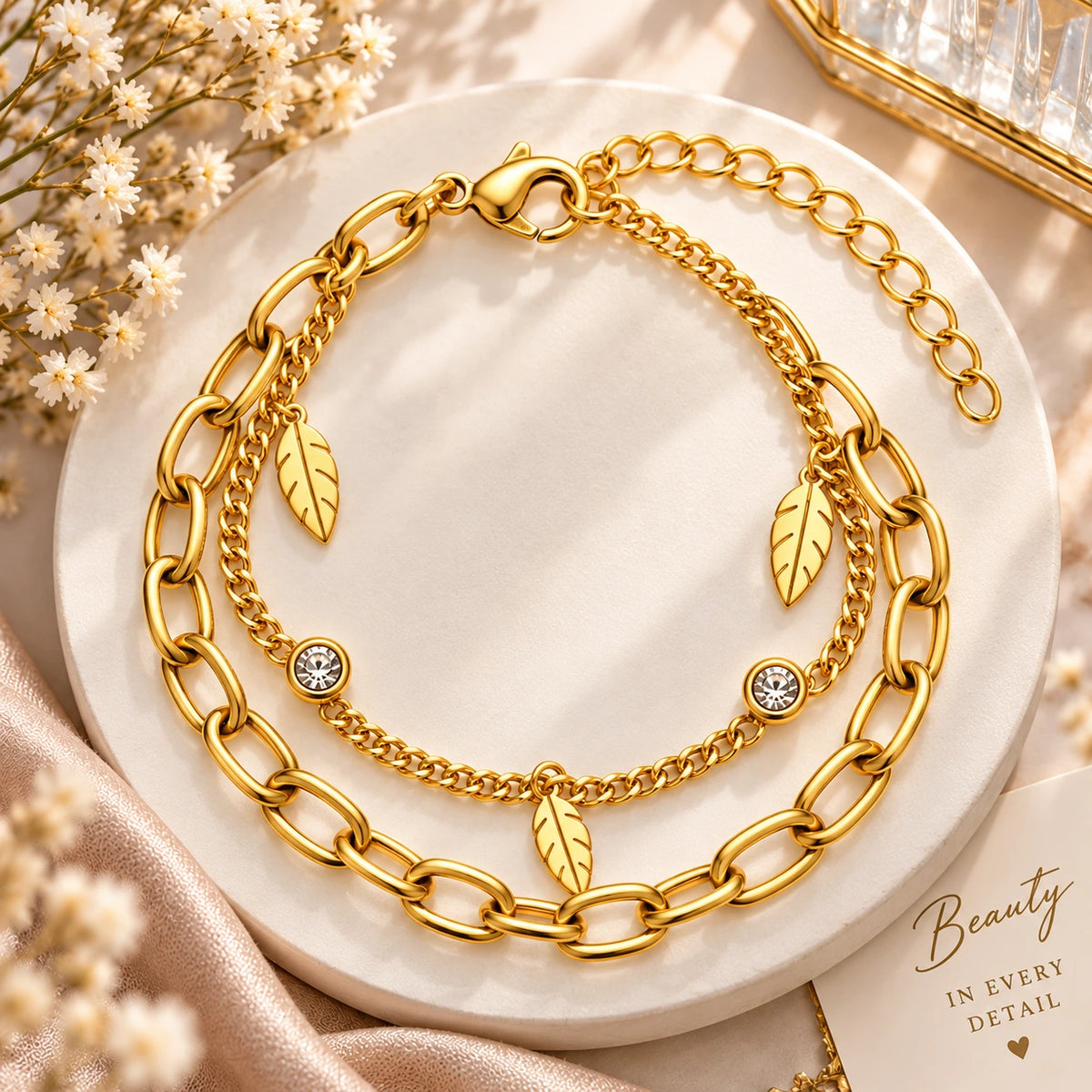 A gold-toned double-layer bracelet with chunky oval links, delicate chain links, leaf charms, and crystal-studded accents, displayed on a round white marble tray with dried flowers and soft beige fabric.