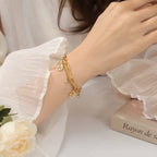 A close-up of a person's wrist wearing a delicate gold chain bracelet with small charms, including a crescent moon and a tiny heart, styled with a white ruffled sleeve and a soft neutral background.
