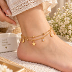 A close-up of a person's ankle wearing a delicate gold double-layer anklet with charms including a star, crescent moon, and a small clear rhinestone, styled with lace fabric and white flowers.