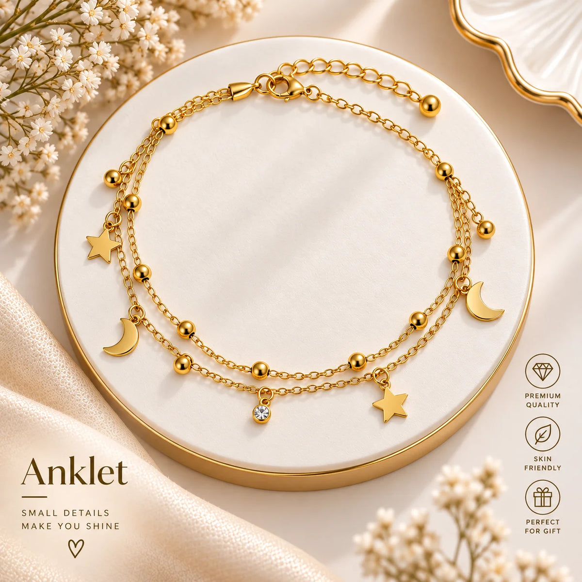 A gold anklet with three layered chains, featuring crescent moon, star, and bead charms, displayed on a round white and gold tray with decorative dried flowers and satin fabric.