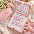 A hand holds an open pink compact mirror displaying pastel press-on nails with pink and lavender colors, white bows, hearts, and plaid patterns. The background includes a pink box, white daisies, and soft pink fabric.
