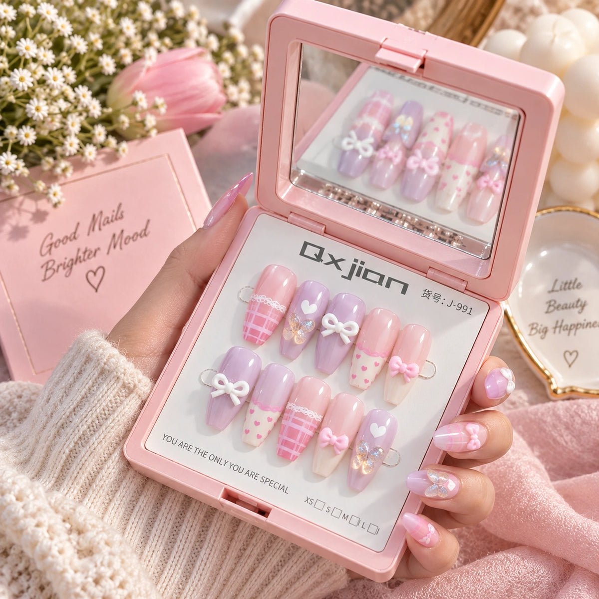 A hand holds an open pink compact mirror displaying pastel press-on nails with pink and lavender colors, white bows, hearts, and plaid patterns. The background includes a pink box, white daisies, and soft pink fabric.