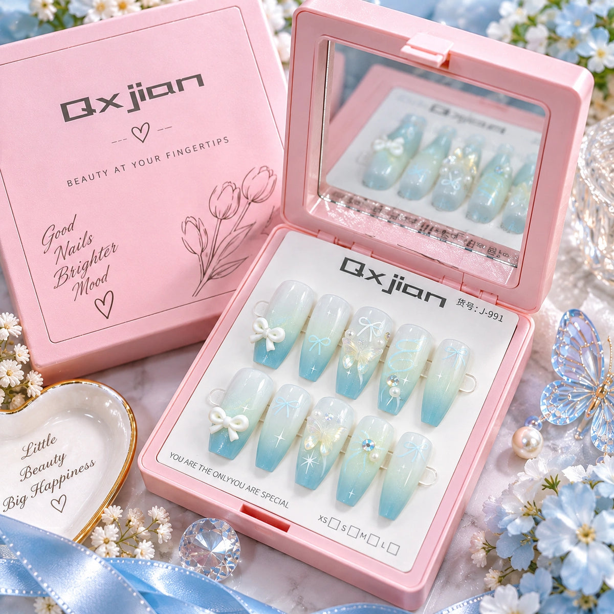 A pink box of Qxjian false press-on nails shown open next to its matching pink lid. The nails are light blue to white ombre with white bows and gold glitter accents.