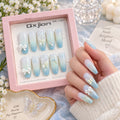 A hand wearing long almond-shaped press-on nails with a light blue to white gradient, decorated with white bows and small iridescent star details, is shown beside an open pink product box labeled Qxjian. The scene includes soft floral props and a decorative tray, making it a styled lifestyle product shot.