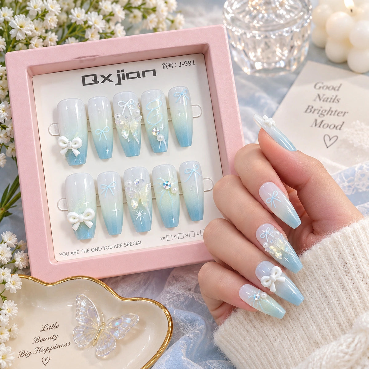 A hand wearing long almond-shaped press-on nails with a light blue to white gradient, decorated with white bows and small iridescent star details, is shown beside an open pink product box labeled Qxjian. The scene includes soft floral props and a decorative tray, making it a styled lifestyle product shot.