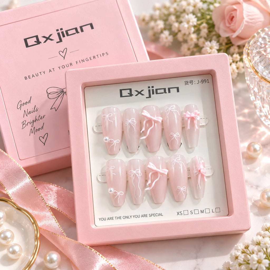 A set of 10 false press-on nails in a pink square box, displayed with the matching pink Qxjian product box and decorative pink ribbons, pearls, and flowers on a light surface.