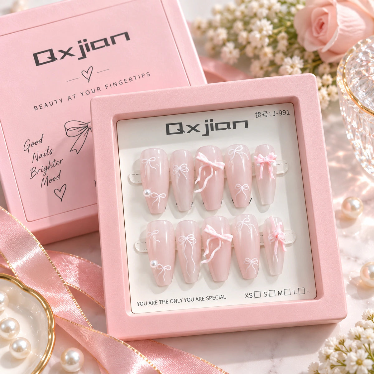 A set of 10 false press-on nails in a pink square box, displayed with the matching pink Qxjian product box and decorative pink ribbons, pearls, and flowers on a light surface.
