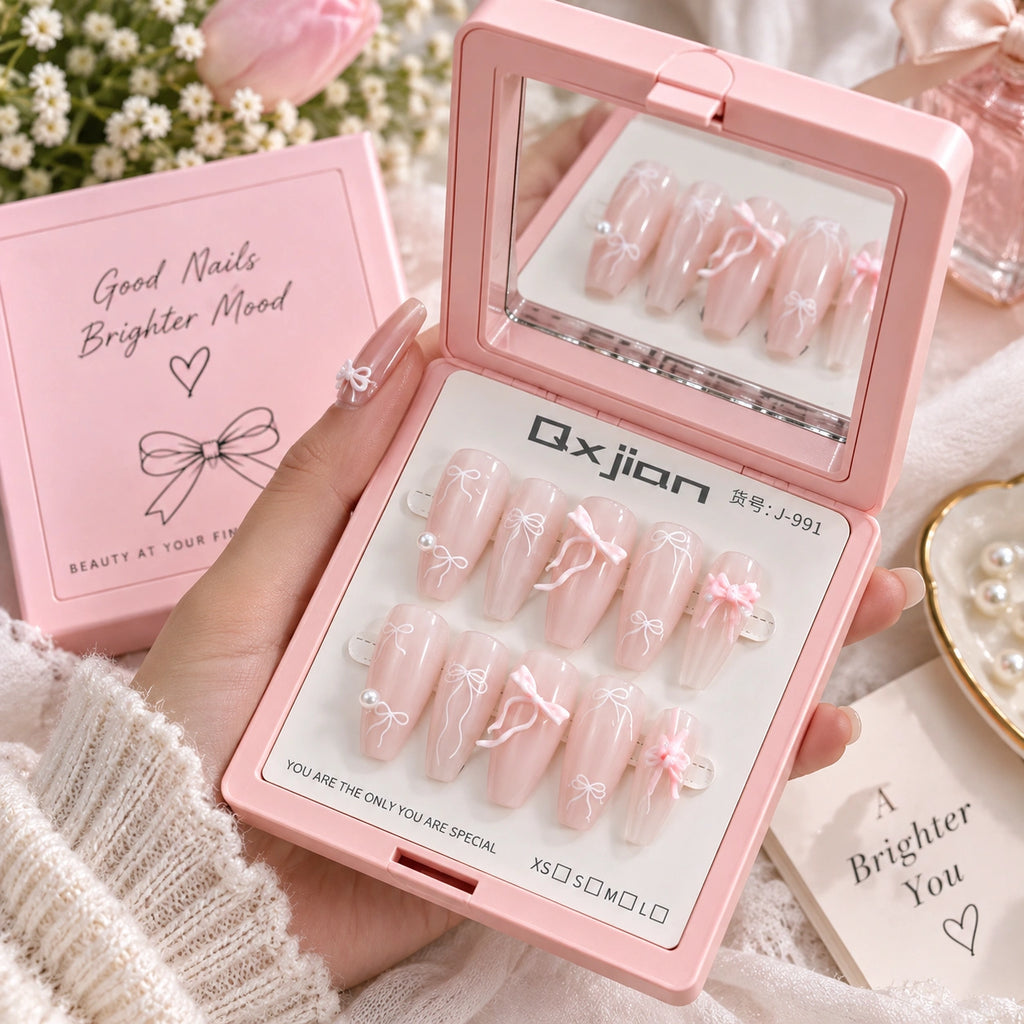 A hand holds an open pink press-on nail kit showing 10 decorated pink nails with white lace and bow designs. The kit includes a mirror on the inside lid, a pink box labeled 'Good Nails Brighter Mood,' and a card reading 'A Brighter You.