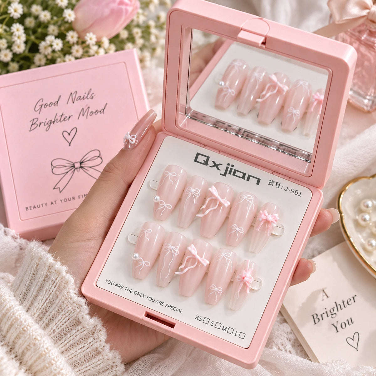 A hand holds an open pink press-on nail kit showing 10 decorated pink nails with white lace and bow designs. The kit includes a mirror on the inside lid, a pink box labeled 'Good Nails Brighter Mood,' and a card reading 'A Brighter You.